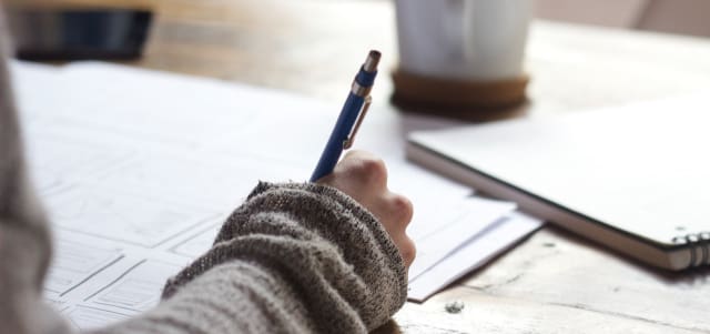 up close shot of someone wearing long sleeves holding a pen writing on a large document next to a notebook on the table