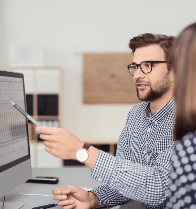 white male in glasses pointing a pen at a computer screen in focus and an out of focus woman listening to him