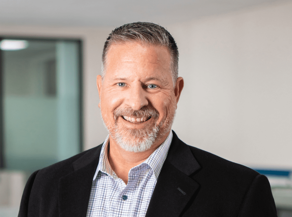 professional headshot of a white male in leadership with a grey beard and smiling in a black suit with office background