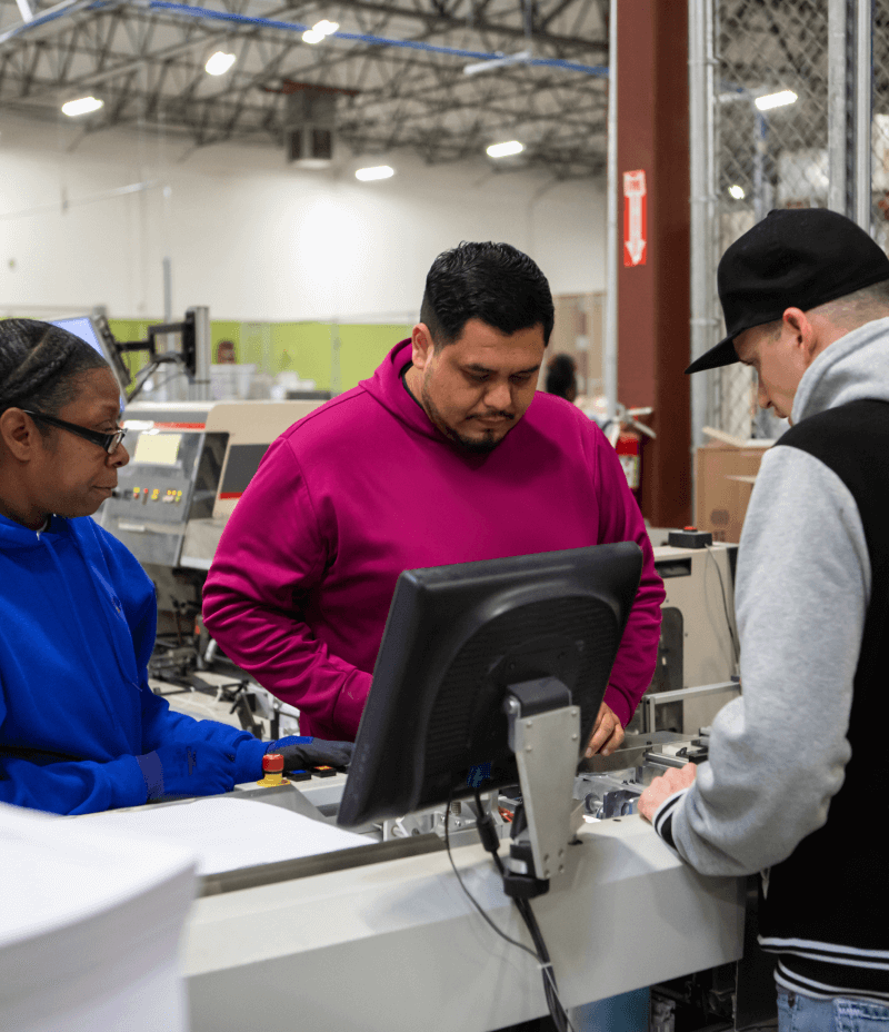 two employees behind a desk in a printing facility helping a customer in a black hat