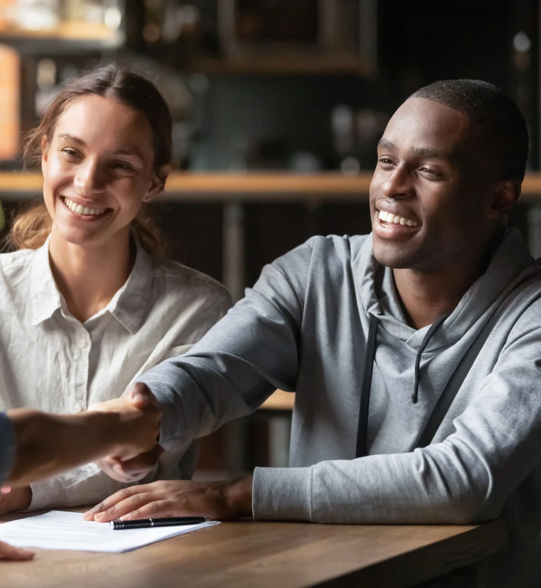 Man and woman smiling as the man shakes hands with another person across a desk