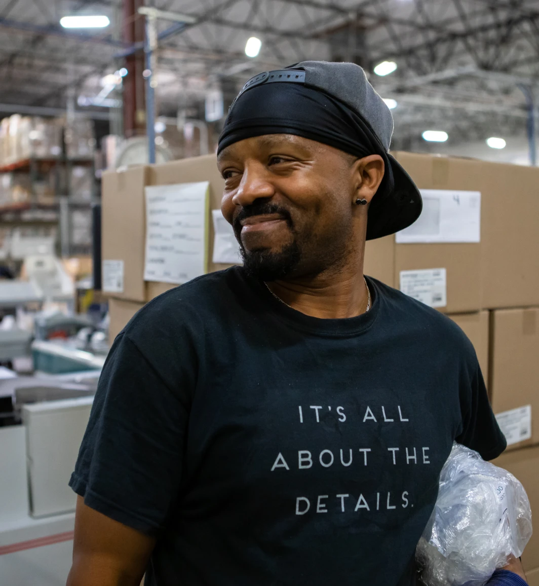 close-up shot of a Black male in a hat and black shirt smiling in a warehouse facility with brown boxes