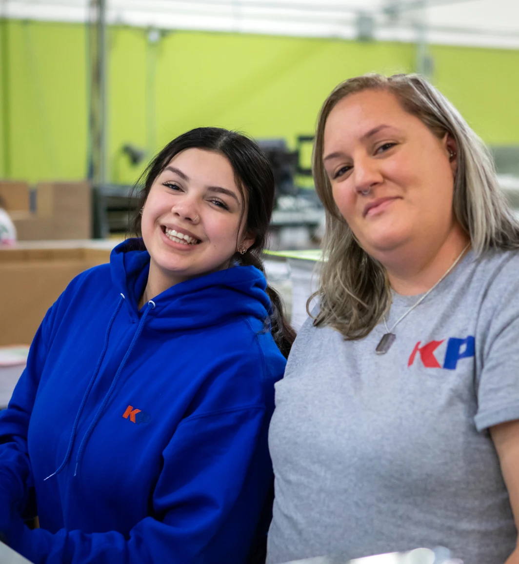 two women smiling one in a KP Corp blue sweatshirt and one in a grey KP Corp short sleeve shirt