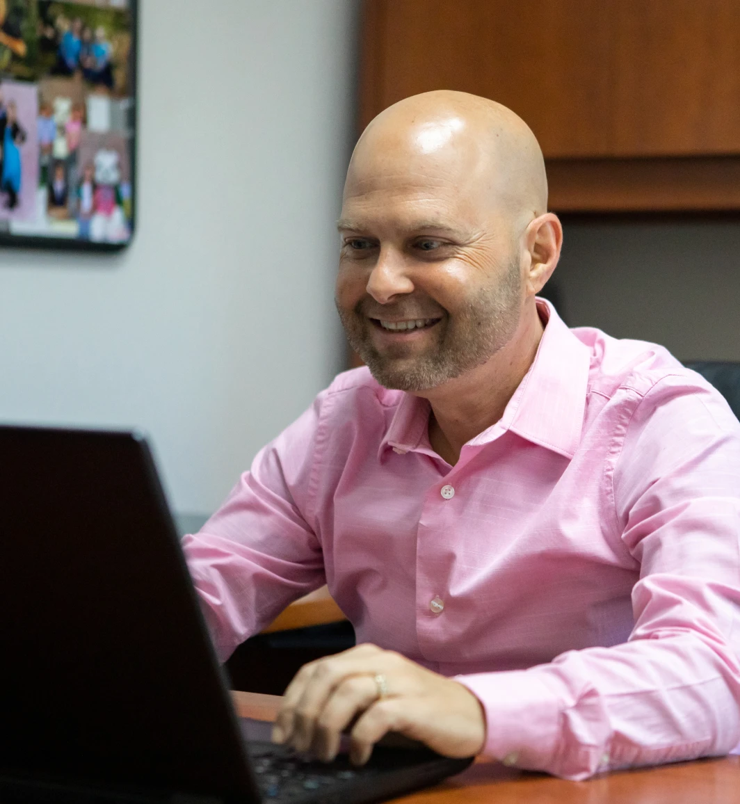 smiling bald white man in a pink button-up collared shirt working on a laptop on a desk