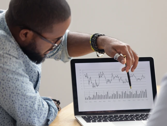 Man gesturing with his pen at a laptop screen showing a graph