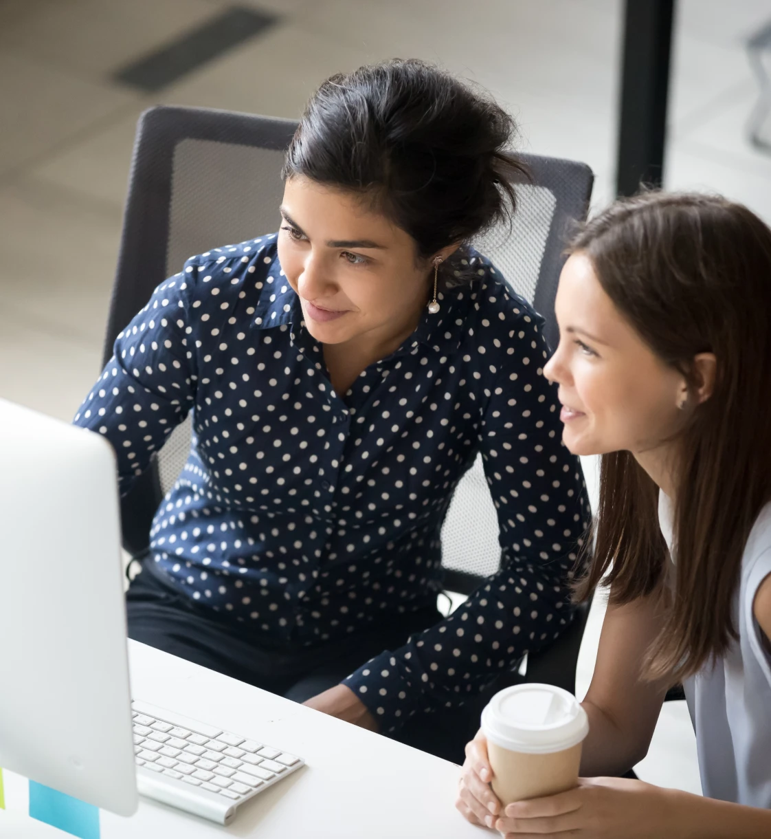 two women drinking a coffee and looking at a computer screen in an organized white office