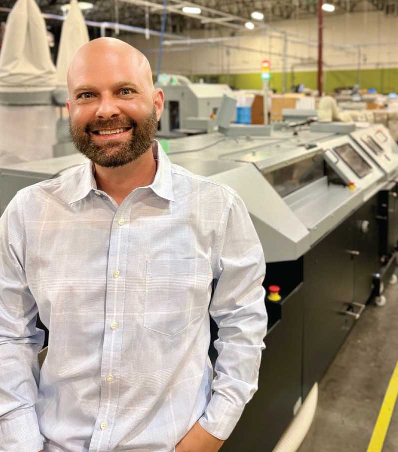 close up of a bald man with a beard smiling and standing in front of a modern printing press machine in a warehouse