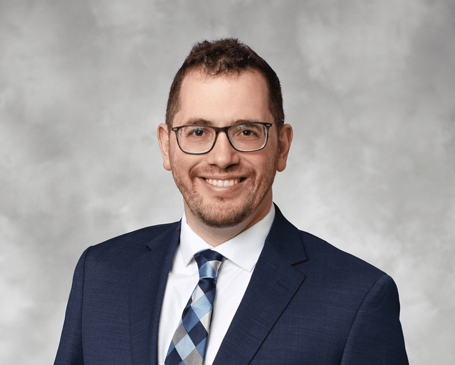 professional headshot of a white male named adam wearing glasses and smiling in a suit with a grey wash background